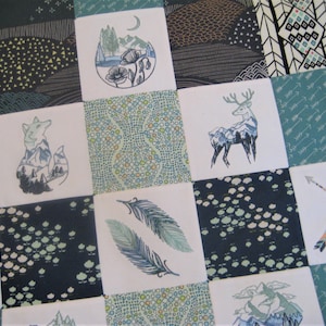 May include: A white fabric quilt with a patchwork design featuring various nature-themed motifs, including a fox, a deer, feathers, and arrows. The quilt is made up of nine squares, each with a different design. The background of the quilt is a dark blue color.