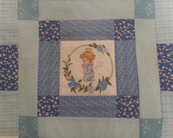 Puede incluir: Una colcha de retazos azul y blanca con una imagen bordada de un ángel rezando en el centro.