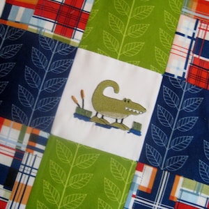 May include: A patchwork quilt with a green, blue, and red plaid pattern. The center square features an embroidered alligator in green.