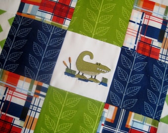 Puede incluir: Una colcha de retazos con un patrón de cuadros verde, azul y rojo. El cuadrado central presenta un cocodrilo bordado en verde.