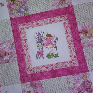 May include: A pink and white patchwork quilt with a central square featuring an embroidered image of two fairies. The fairies are surrounded by flowers and leaves.
