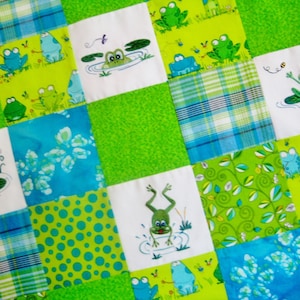 May include: A green and blue patchwork quilt with a frog theme. The quilt is made of various fabrics, including plaid, polka dots, and floral prints. The frogs are embroidered in different poses, some with lily pads and others with bees.