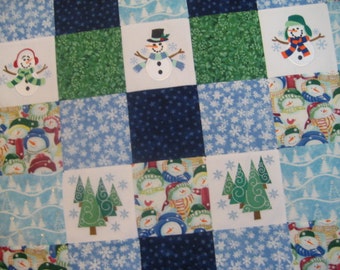 Könnte beinhalten: Eine Patchwork-Decke mit Winterthema. Die Decke besteht aus verschiedenen blauen, grünen und weißen Stoffen. Jedes Quadrat zeigt ein gesticktes Design eines Schneemanns oder einer Gruppe von Schneemännern. Einige Quadrate haben ein Schneeflockenmuster.