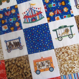 May include: A colorful patchwork quilt with various embroidered designs, including a circus tent, animals in cages, popcorn, and peanuts. The quilt is made of white fabric with blue, red, and yellow accents.