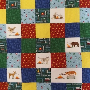 May include: A colorful quilt with a camping theme. The quilt is made of squares of different fabrics, each with a different design. Some of the designs include a campfire, a tent, a bear, a deer, a wolf, and a fox. The quilt also features the words "campfire", "tent", "moonlight", "fireflies", and "s'mores".