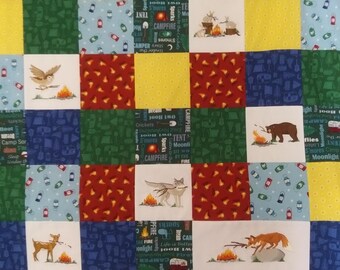 Puede incluir: Una colcha colorida con un tema de campamento. La colcha está hecha de cuadrados de diferentes telas, cada uno con un diseño diferente. Algunos de los diseños incluyen una fogata, una tienda de campaña, un oso, un ciervo, un lobo y un zorro. La colcha también presenta las palabras "fogata", "tienda de campaña", "luz de luna", "luciérnagas" y "malvaviscos".