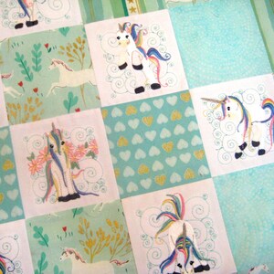 May include: A patchwork quilt with a light blue background and white squares featuring embroidered unicorns with rainbow manes and tails. The squares are surrounded by a green border with gold accents.