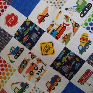 May include: A colorful patchwork quilt with a variety of construction vehicle designs. The quilt features a stop sign, road work sign, and under construction sign. The quilt is made of different fabrics, including blue, red, white, and yellow.
