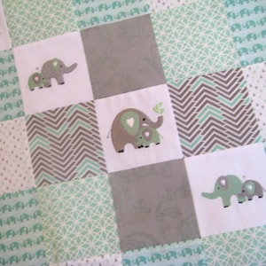 May include: A baby quilt with a patchwork design featuring various shades of green and gray. The quilt is made of different fabrics with various patterns, including a chevron pattern, a geometric pattern, and a pattern of small elephants. The quilt features embroidered elephants in various sizes.