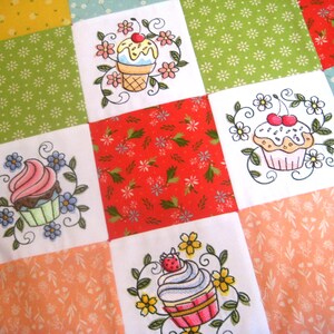 May include: A white fabric quilt with a patchwork design featuring embroidered cupcakes with different colored frosting and toppings. The quilt is made up of squares of different colors and patterns, including red, green, yellow, and pink.