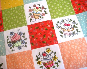 Puede incluir: Una colcha de tela blanca con un diseño de patchwork que presenta cupcakes bordados con glaseados y coberturas de diferentes colores. La colcha está hecha de cuadrados de diferentes colores y patrones, incluyendo rojo, verde, amarillo y rosa.