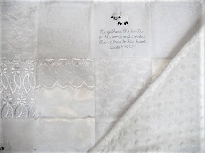 K&ouml;nnte beinhalten: Wei&szlig;e Babydecke mit Patchwork-Muster. Die Decke zeigt eine wei&szlig;e Lamm-Applikation und ein Zitat aus der Bibel: "Er sammelt die L&auml;mmer in seine Arme und tr&auml;gt sie nahe an sein Herz. Jesaja 40:11"