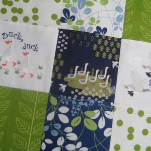 May include: A white fabric quilt with a patchwork design. The quilt is made up of squares with different patterns, including green leaves, blue flowers, and white polka dots. The squares also feature embroidered images of ducks and geese with the text "Duck, duck" and "goose!"