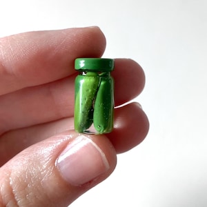 May include: A miniature glass jar with a green lid, containing two small, bright green pickles. The jar is held between fingers against a white background, showcasing the tiny food item.