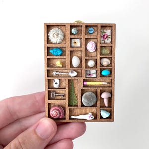 May include: A rectangular wooden shadow box display case featuring multiple small compartments. Each cubby contains miniature objects including a white flower, blue button, playing card, silver spoon, key, pencil, shell, and various stones.
