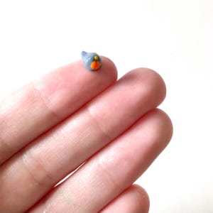 May include: A tiny, gray bird figurine with an orange beak and eye, held between fingers. The miniature sculpture is a detailed representation of a bird, perfect for crafts or collecting. The bird is approximately 0.5 inches in length.