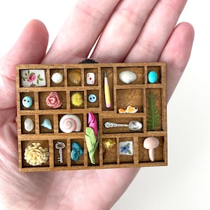 May include: A rectangular wooden shadow box with multiple compartments containing miniature objects including a tiny pencil, a silver spoon, a seashell, a key, a pink flower, a fern leaf, a mushroom, and various small beads and fabric scraps.