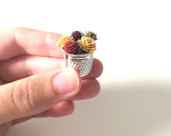 May include: A small silver thimble filled with miniature faux flowers in shades of yellow, orange, and red.