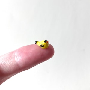 May include: A tiny, yellow ceramic bird figurine with black and orange details. The miniature bird is perched on a fingertip, showcasing its small size and intricate design. The bird's body is yellow with black markings, and it has a black head and orange beak.