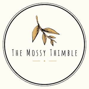 May include: A white circle with a black outline and a gold branch with leaves in the center. The text "THE MOSSY THIMBLE" is written below the branch.