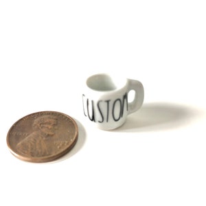 May include: A small white ceramic mug with a black "CUSTOM" inscription. The mug is next to a US penny for scale.