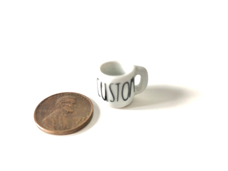 May include: A small white ceramic mug with a black "CUSTOM" inscription. The mug is next to a US penny for scale.