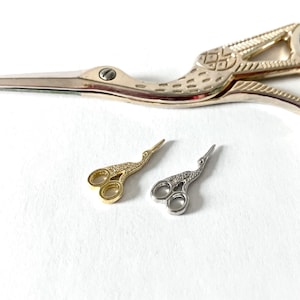 May include: A gold-colored and silver-colored pair of small, decorative stork scissors, with a larger pair of gold scissors in the background. The scissors are on a white surface.