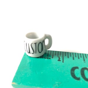 May include: A small white ceramic mug with a black "CUSTOM" inscription. The mug is sitting on a green ruler.