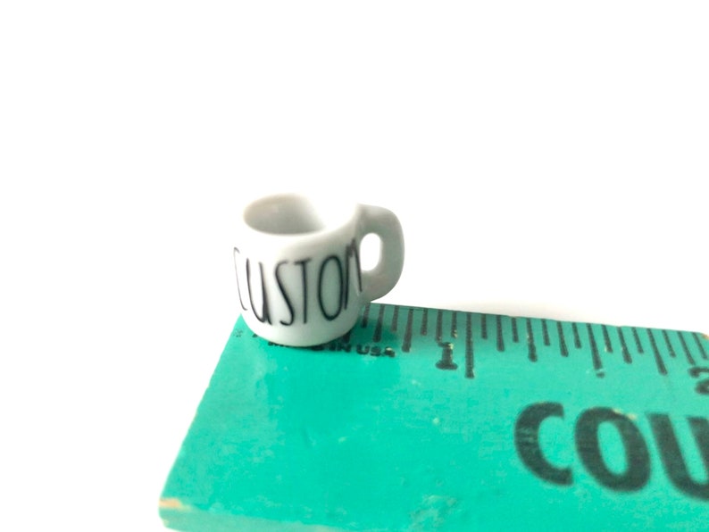 May include: A small white ceramic mug with a black "CUSTOM" inscription. The mug is sitting on a green ruler.