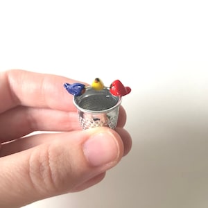 May include: A silver thimble with three small ceramic birds perched on the rim. The birds are blue, yellow, and red.