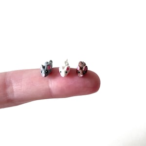 May include: Three miniature mice figurines, one gray, one white, and one brown, are lined up on a finger. The mice have pink ears and are facing forward.