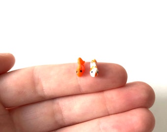 May include: Two small ceramic goldfish figurines, one orange and one white with orange spots, are displayed on a finger.