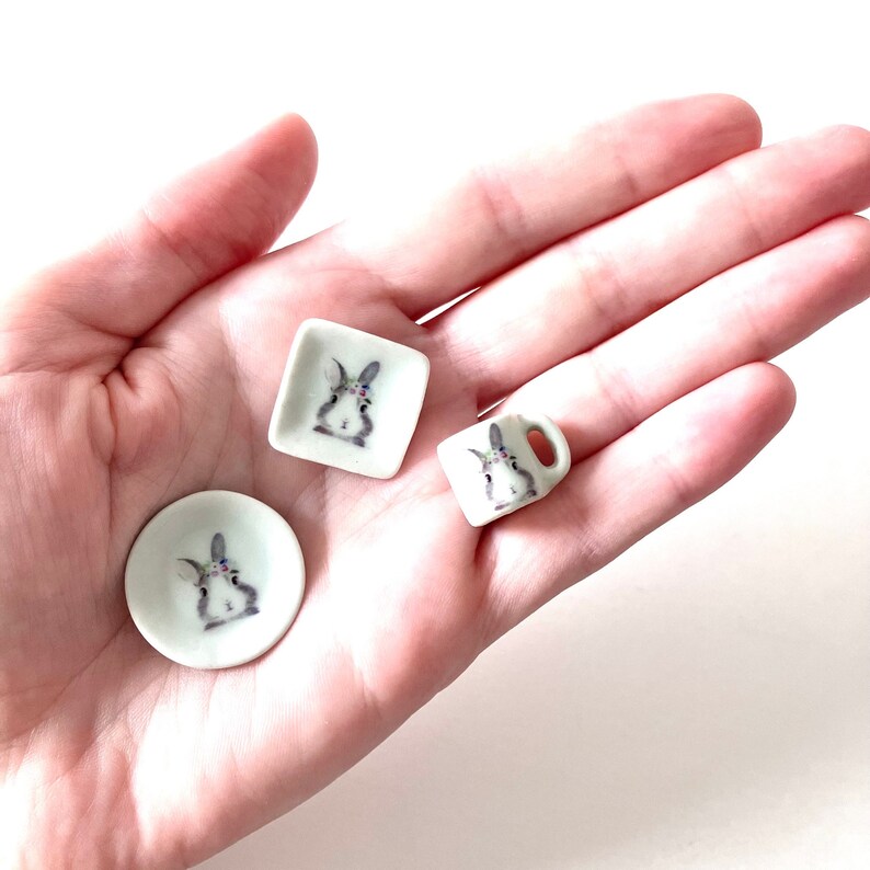 May include: A miniature tea set with a bunny design. The set includes a small square plate, a round plate, and a cup with a handle. The bunny is depicted in a light gray color with a floral crown.