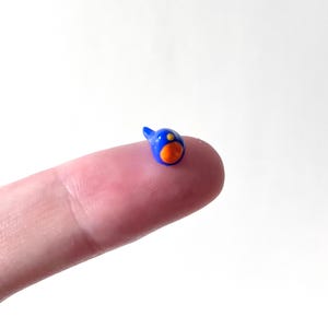May include: A tiny, blue bird figurine with an orange belly and a yellow dot on its head. The miniature bird has a small, pointed beak and is perched on a fingertip against a white background. The figurine is likely made of ceramic or glass.