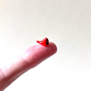 May include: A tiny, red, chili pepper-shaped glass bead with a black dot, resting on a fingertip. The bead is glossy and appears to be handmade. The background is a plain, off-white color.