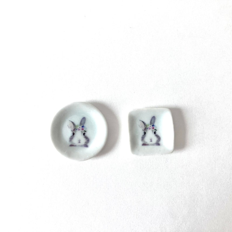 May include: Two white ceramic plates with a bunny rabbit design. The larger plate is round and the smaller plate is square. Both plates feature a gray bunny with a floral headband.