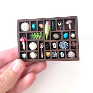 May include: A miniature wooden display case filled with various small objects. The case contains a variety of items, including a mushroom, a key, a spoon, a seashell, and a playing card. The objects are arranged in individual compartments.