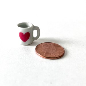 May include: A small white ceramic mug with a pink heart design. The mug is about the size of a US penny.