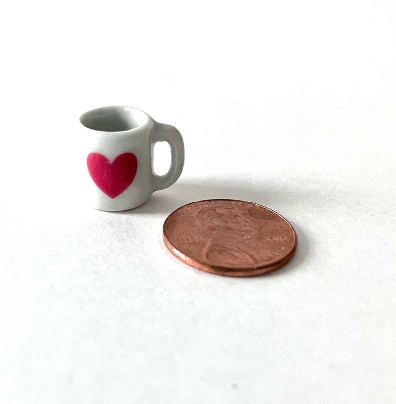 May include: A small white ceramic mug with a pink heart design. The mug is about the size of a US penny.