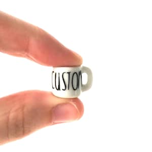 May include: A small white ceramic mug with a black "CUSTOM" inscription. The mug is being held by a hand.