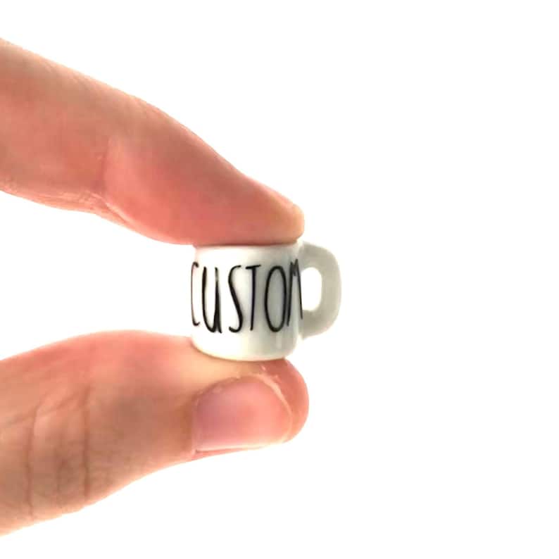 May include: A small white ceramic mug with a black "CUSTOM" inscription. The mug is being held by a hand.