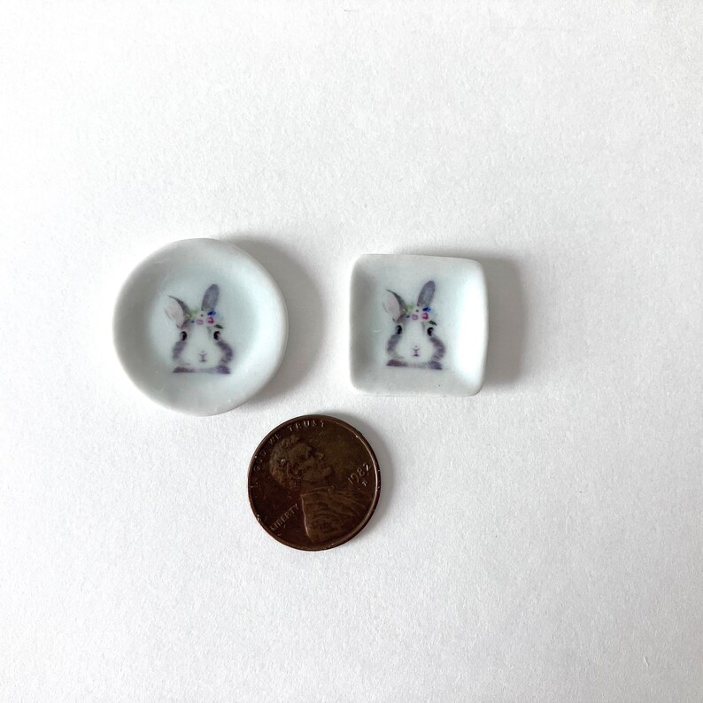 May include: Two miniature ceramic plates with a bunny rabbit design. The plates are white with a gray bunny rabbit wearing a flower crown. The plates are about the size of a US penny.