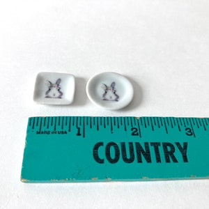 May include: Two small white ceramic plates with a bunny rabbit design. The plates are about 1 inch in diameter and are sitting on a green ruler.