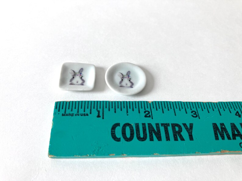 May include: Two small white ceramic plates with a bunny rabbit design. The plates are about 1 inch in diameter and are sitting on a green ruler.