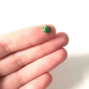 May include: A small green frog figurine, about the size of a fingernail, is held in a person's hand.
