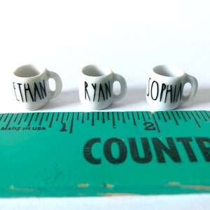 May include: Three white ceramic mugs with black lettering. The mugs are labeled "Ethan", "Ryan", and "Sophia".