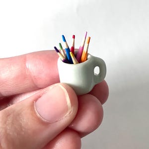 May include: A miniature white ceramic mug filled with colorful pencils. The pencils have various colors, including blue, green, red, and orange. The mug has a small handle and is held in a person's hand. The pencils are arranged upright.