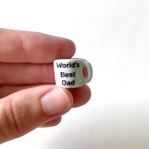May include: A small, light green mug with a handle, held between fingers. The mug has the words "World's Best Dad" printed in black. The mug is a miniature size.