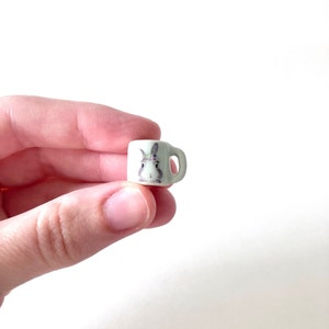 May include: A tiny white ceramic mug with a bunny rabbit design. The mug has a small handle and is about the size of a fingertip.