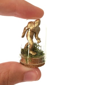 May include: A gold-colored Bigfoot figurine is displayed inside a clear glass jar with a cork stopper. The jar is filled with green moss.
